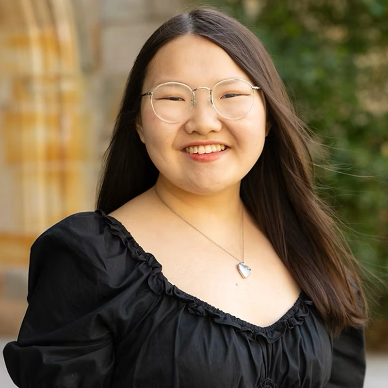 Estelle Balsirow, a young woman with long straight dark hair, wearing glasses, a silver heart necklace, and a black ruffled dress, smiles outdoors against blurred green foliage and a tan stone building.