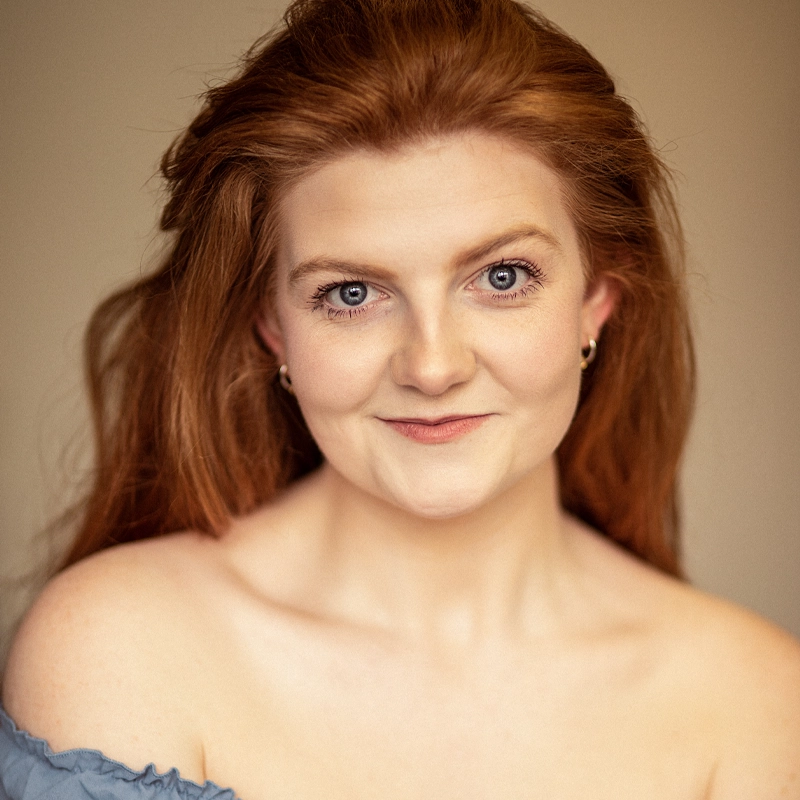 Emeline Battles, with long red hair and fair skin, smiles softly at the camera. She wears small hoop earrings and an off-the-shoulder blue top. The neutral, blurred background highlights her expressive eyes and gentle expression.
