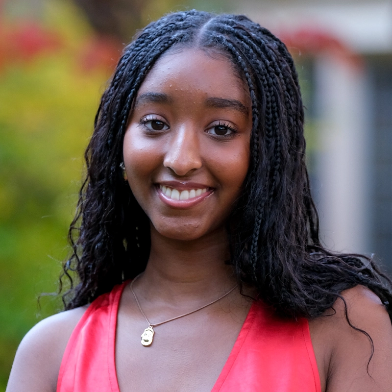 A young woman with dark, braided hair smiles at the camera. She wears a sleeveless red top and a gold necklace with a small pendant. The background is blurred, showing green and yellow foliage, suggesting an outdoor setting.