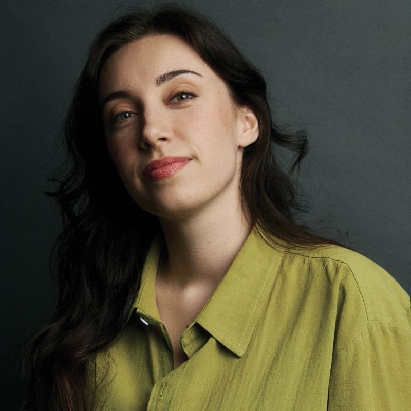 A young woman with long, dark wavy hair and a subtle smile poses against a dark gray background. She is wearing a button-down shirt in a muted chartreuse color. Light hits her face from the side, highlighting her features.