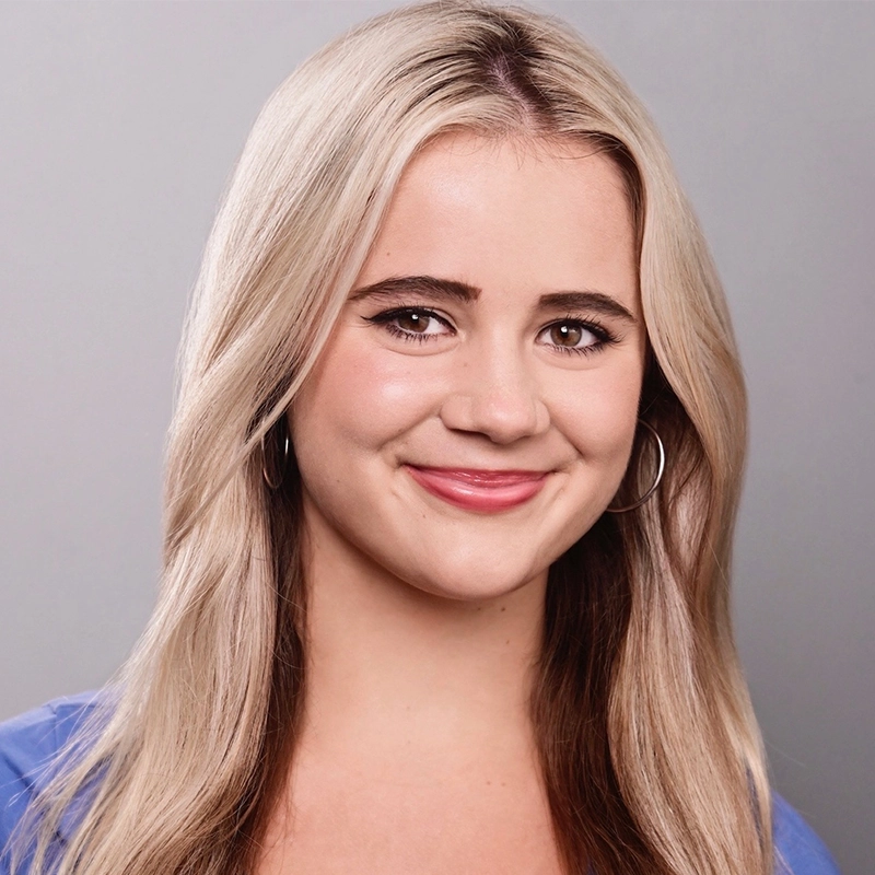 A young woman with long, straight blonde hair and fair skin smiles at the camera. She has brown eyes, wears silver hoop earrings, and a blue top. The background is plain light gray, giving the image a professional headshot style.