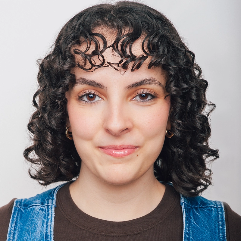 Sara Ross Avery, a young woman with short, dark curly hair and defined baby bangs, smiles slightly. She wears small gold hoop earrings, a brown shirt, and a blue denim overall. Her blue eyes stand out as she poses in front of a plain, light background.
