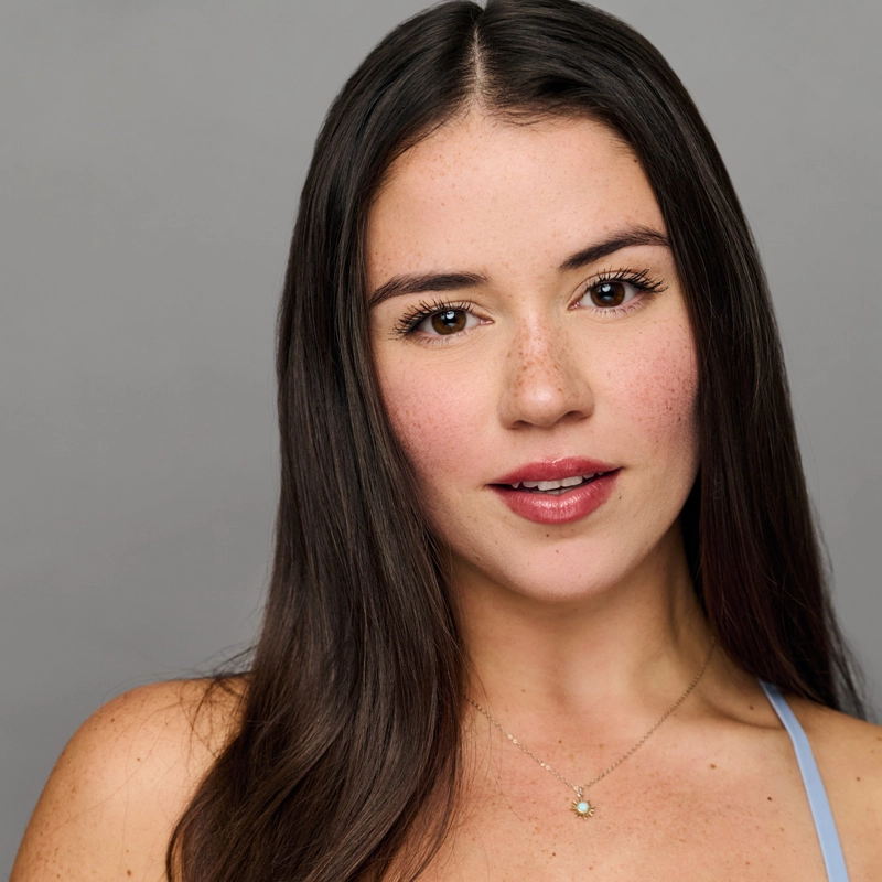 Allie Beltran, a young woman with long, straight dark brown hair and fair skin, looks at the camera. She has natural makeup, pink lips, freckles on her cheeks, and wears a delicate necklace with a light blue top against a gray background.