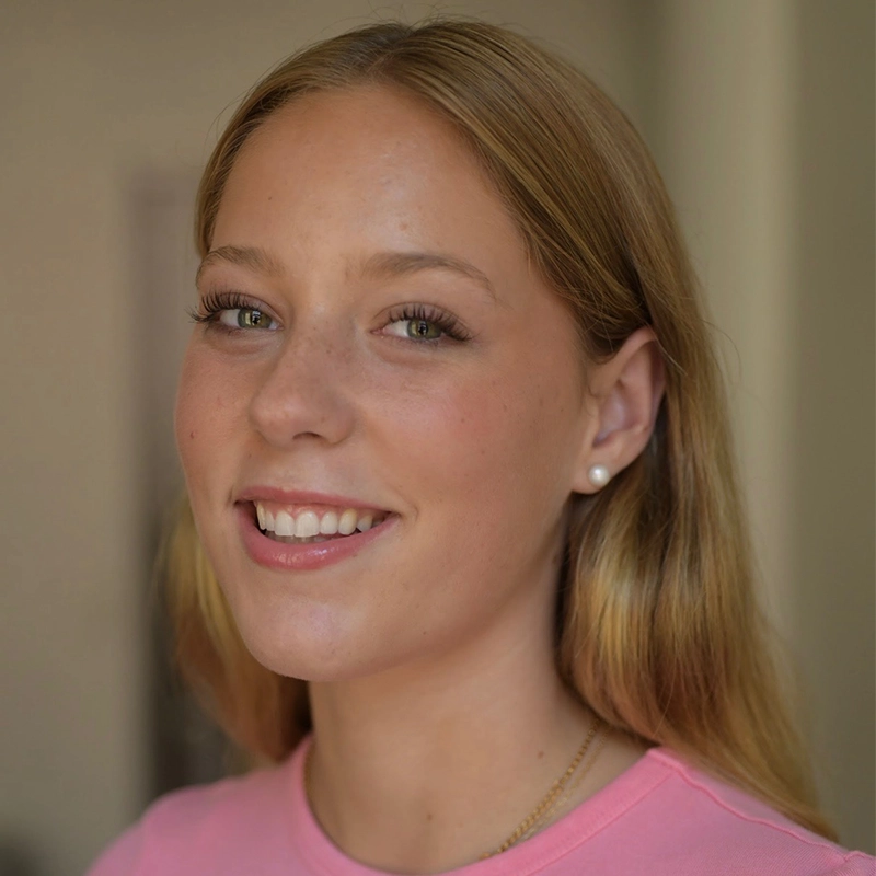 A young woman with long blonde hair smiles softly. She wears a pink top, a gold necklace, and pearl stud earrings. The softly blurred background draws focus to her calm, friendly face—an inviting portrait by Micah Wolfensohn.