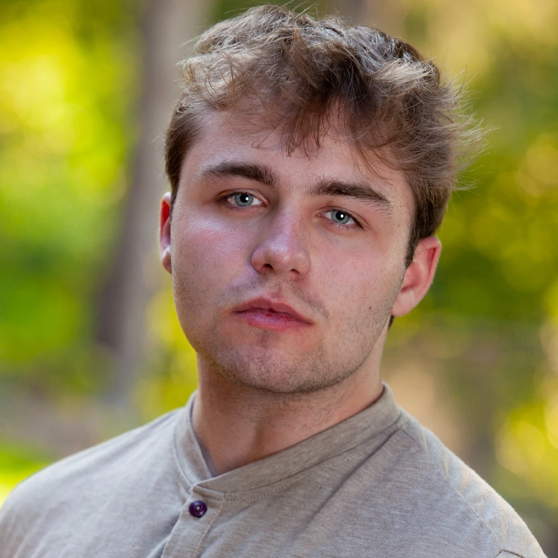 Nicholas Bordoni, a young man with tousled light brown hair and blue eyes, looks seriously at the camera. He wears a beige buttoned shirt, while the blurred green background suggests an outdoor setting with trees and sunlight.