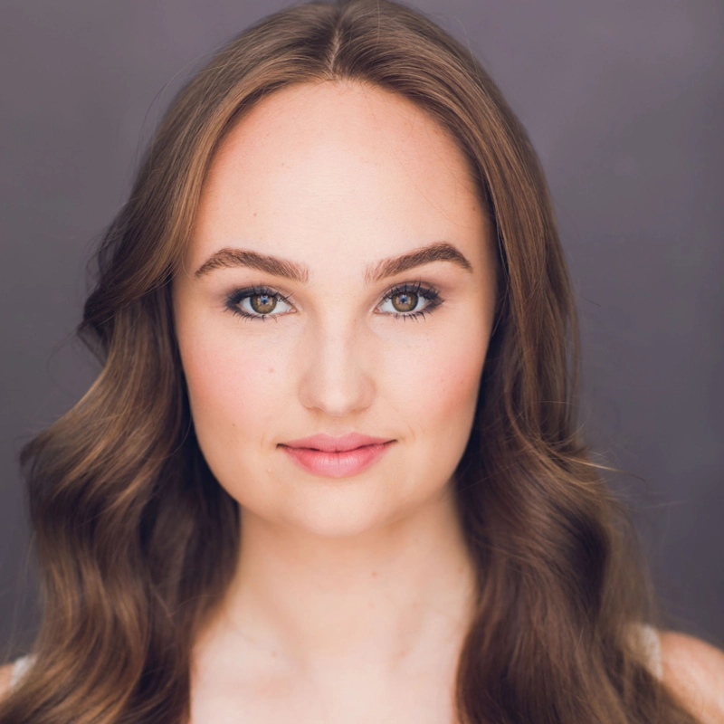 A young woman with long, wavy brown hair and fair skin looks directly at the camera. She has arched eyebrows, light hazel eyes, and a soft pink lip color. The background is plain grey, and the lighting is even, highlighting her facial features.