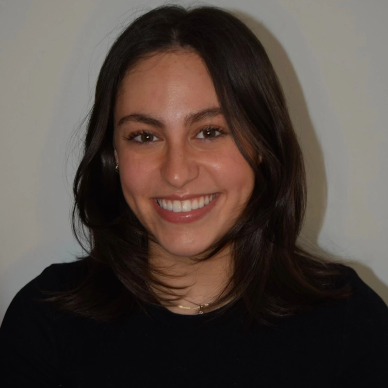 Morgan Brandt, a young woman with straight, shoulder-length brown hair, smiles at the camera. She has light skin, dark eyes, and wears a black top with a thin necklace. The plain, light-colored background creates a warm portrait.
