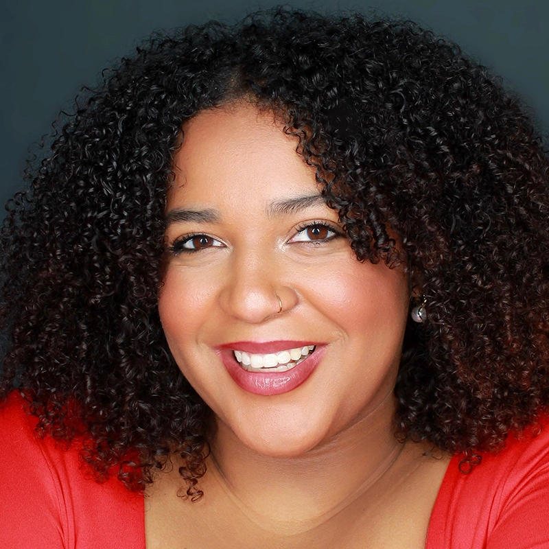 A woman with medium-dark skin and curly dark hair smiles at the camera. She wears a red top, silver stud earrings, a nose ring, and natural makeup with pink lipstick. The background is dark and plain, highlighting her face.