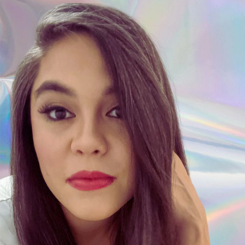 A woman with long, straight dark hair and bold red lipstick gazes at the camera. She has full eyebrows, dark eye makeup, and wears a white top. Her hand rests near her head. The iridescent pastel background adds a dreamy Brisa effect.