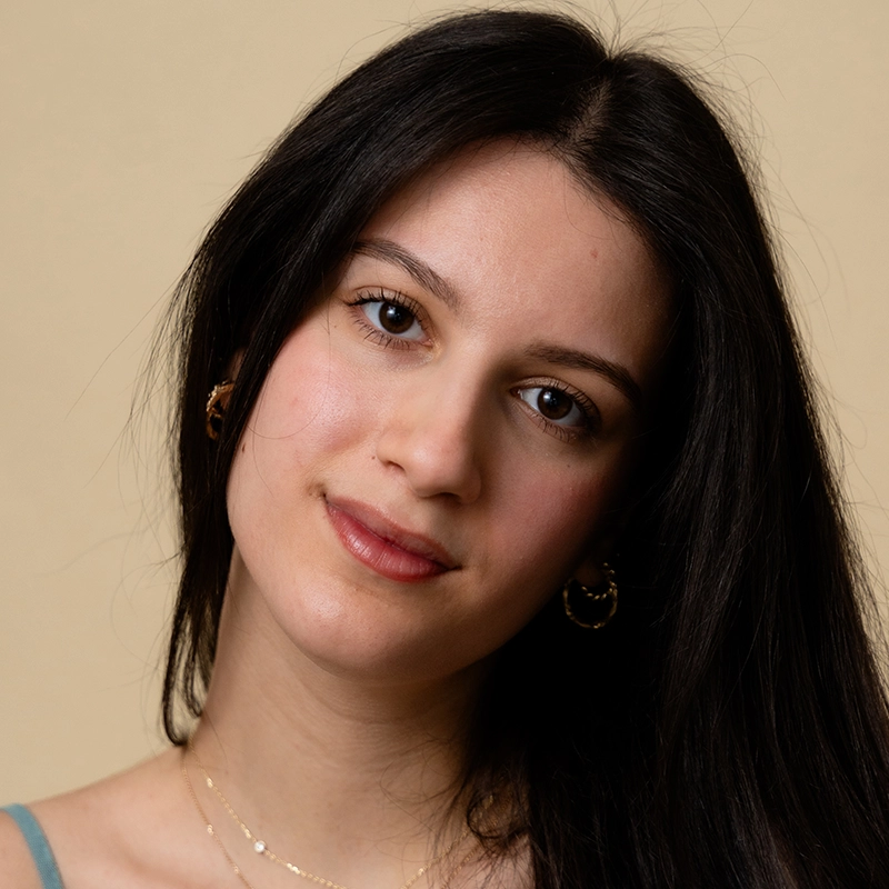 A young woman with long, straight dark hair and light skin looks at the camera with a gentle expression. She is wearing small hoop earrings, a thin strap top, and delicate layered necklaces—her style reminiscent of Lazarus Rashid’s understated elegance.