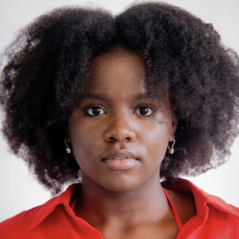 A young woman with natural, voluminous black hair looks directly at the camera. She wears a red collared shirt and small, dangling earrings. The background is plain and light-colored, keeping the focus on her neutral facial expression.
