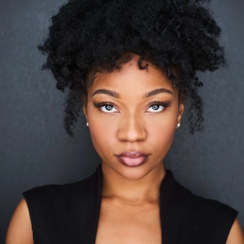 A woman with deep brown skin, bright blue eyes, and full lips stares directly at the camera. Seania Burnett's voluminous, curly black hair is styled in a natural afro updo, and she wears a sleeveless black top against a dark gray background.