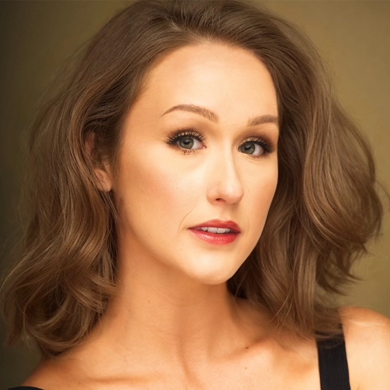 A woman with wavy, shoulder-length brown hair looks at the camera. She has fair skin, light eye makeup, and red lipstick. She is wearing a black top with thin straps against a softly lit, neutral background.