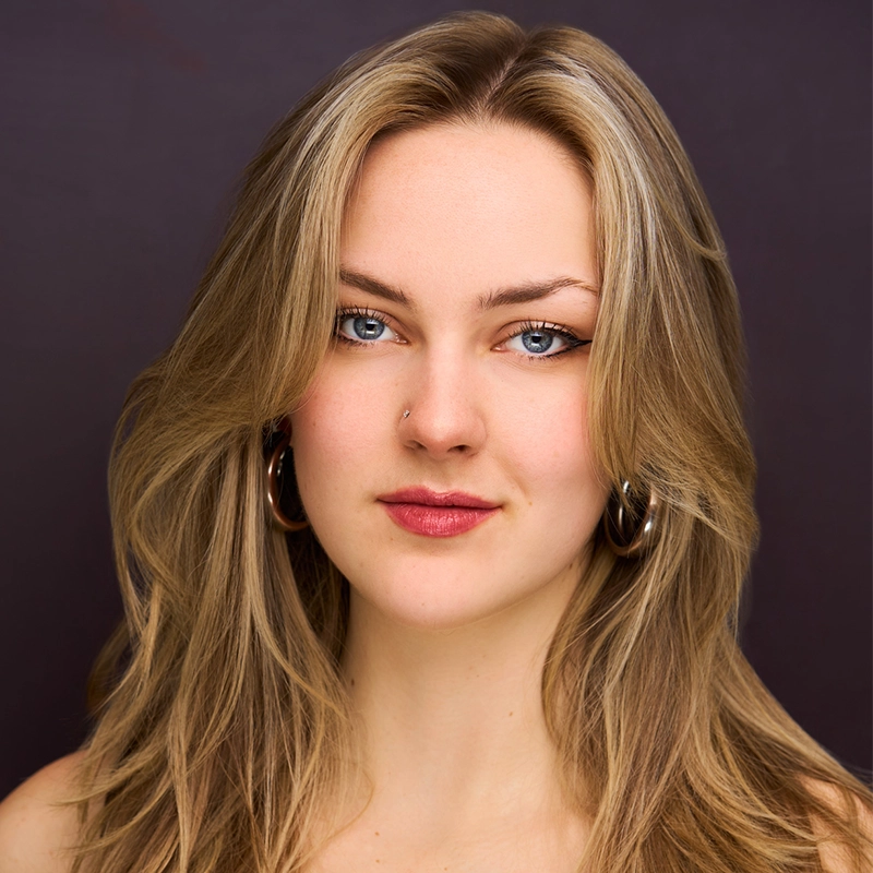 A young woman, Addison Zacharias, with long, layered blonde hair and blue eyes looks confidently at the camera. She has subtle makeup, a small nose piercing, and wears large hoop earrings. The plain dark background highlights her face.