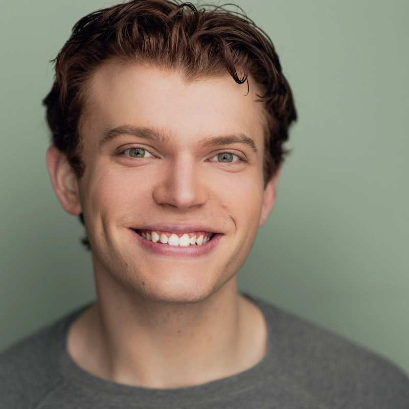 Ben Allen Carter, a young man with light skin and wavy brown hair, smiles warmly at the camera. He has green eyes, visible dimples, and is wearing a grey crew-neck shirt against a solid, muted green background.