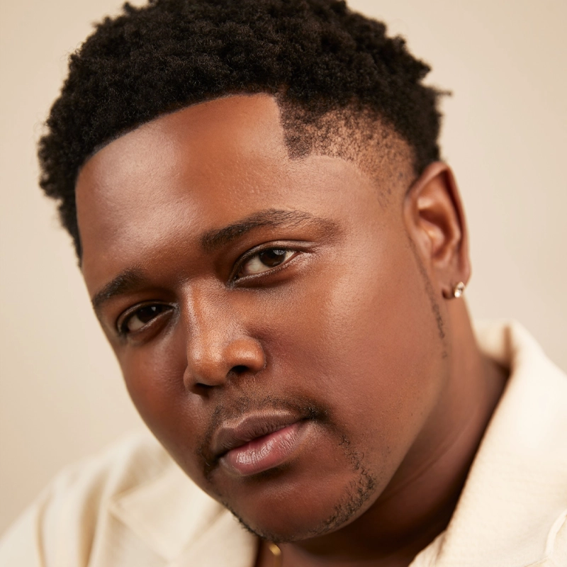 A close-up portrait of Demie Carter, a young man with short curly hair and smooth skin, wearing a cream-colored collared shirt and a small hoop earring. He looks directly at the camera with a calm, confident expression against a neutral background.