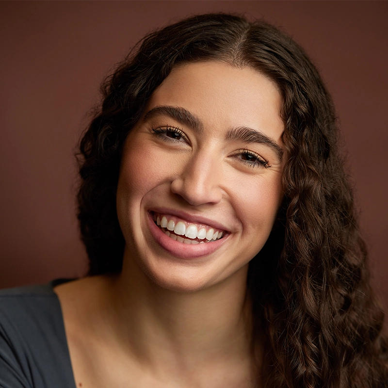 A young woman with long, dark, curly hair smiles warmly at the camera. She has light skin, full eyebrows, and is wearing a gray top. The background is a solid, muted brown, creating a soft and welcoming portrait.