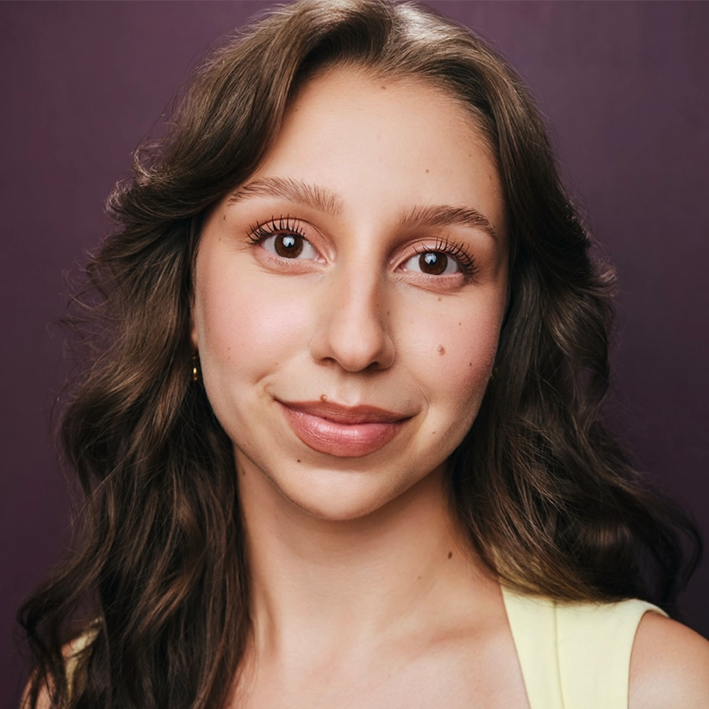 A young woman with long, wavy brown hair and brown eyes smiles softly. She has clear skin, subtle makeup, and is wearing a sleeveless, light yellow top. The background is a plain, deep purple color.