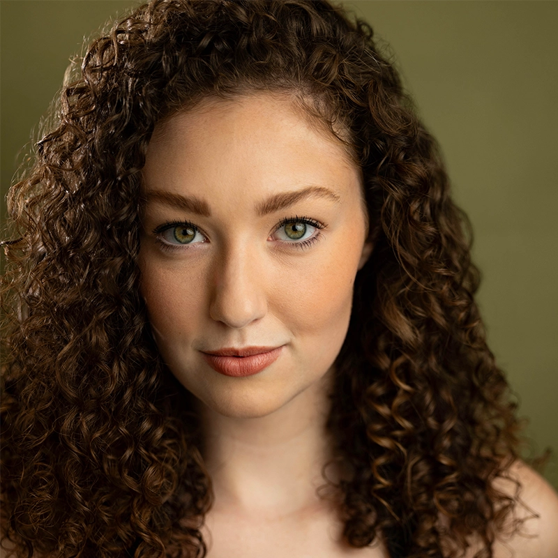 Jenna Willet, a young woman with fair skin and long, thick, brown curly hair, looks directly at the camera. She has green eyes, defined brows, and wears natural makeup with peach-toned lipstick against a muted olive green background.