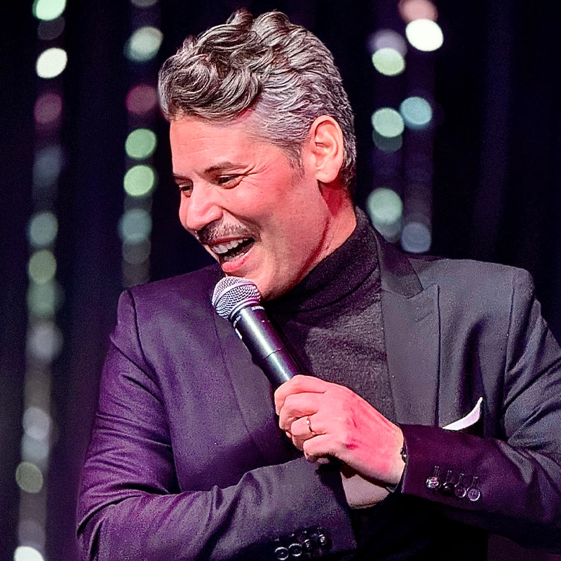 A man with wavy gray hair and a mustache, wearing a dark suit and black turtleneck, smiles and holds a microphone on stage. Joshua Chachi stands before an out-of-focus backdrop with vertical strings of lights, creating a festive atmosphere.
