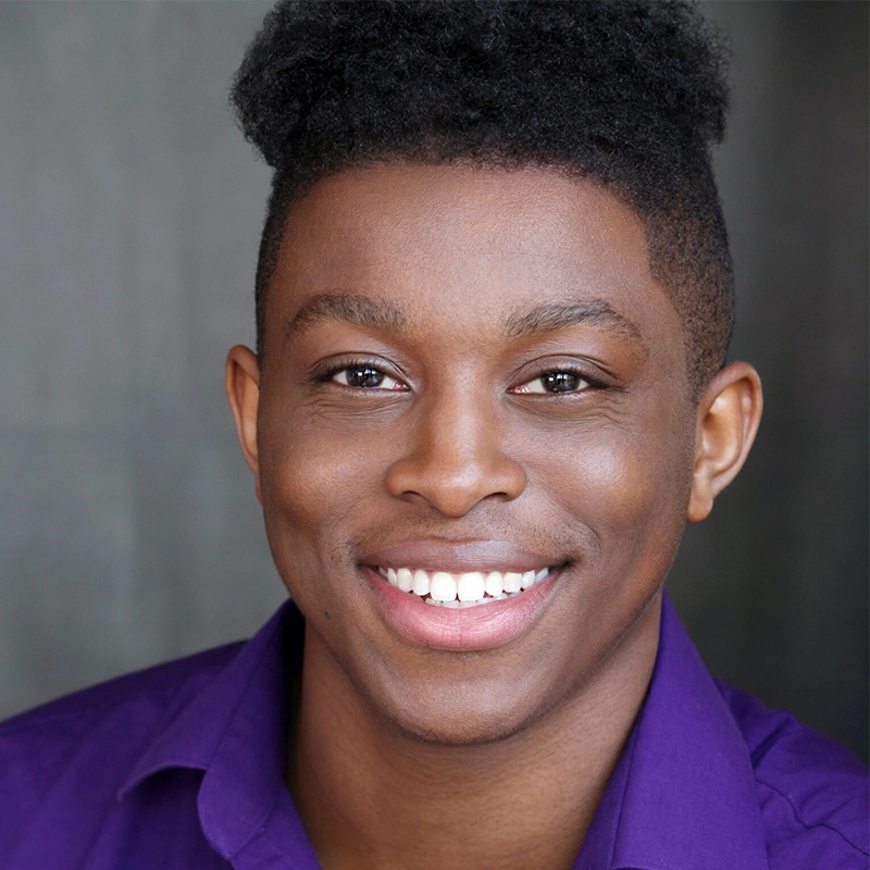 A young person with dark skin and short, curly black hair smiles warmly at the camera. Lexis Trechak wears a purple collared shirt, and the softly blurred gray background keeps focus on their cheerful expression.