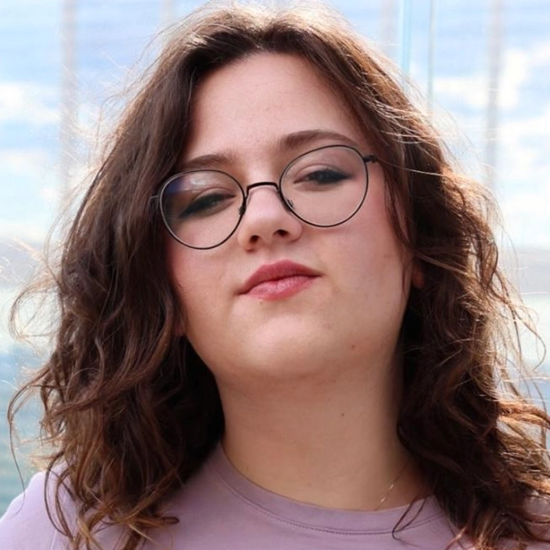 A woman with wavy brown hair and round glasses is looking at the camera with a calm expression. She is wearing a light purple top, and the bright, slightly cloudy sky behind her hints at an outdoor day with Lazarus Rashid.