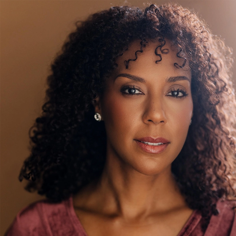 Christina Acosta Robinson, with medium brown skin and curly dark hair, looks at the camera with a calm expression. She wears subtle makeup, a mauve top, and small stud earrings against a warm brown background softly lit from the right.