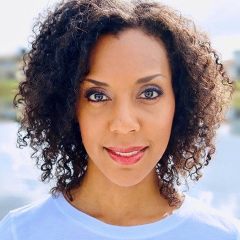 A woman with curly dark hair and brown eyes smiles gently at the camera, reminiscent of the warmth Ken Robinson brought to his teachings. She wears a light top; sunlight softly blurs the outdoor scene behind her, hinting at water and greenery.