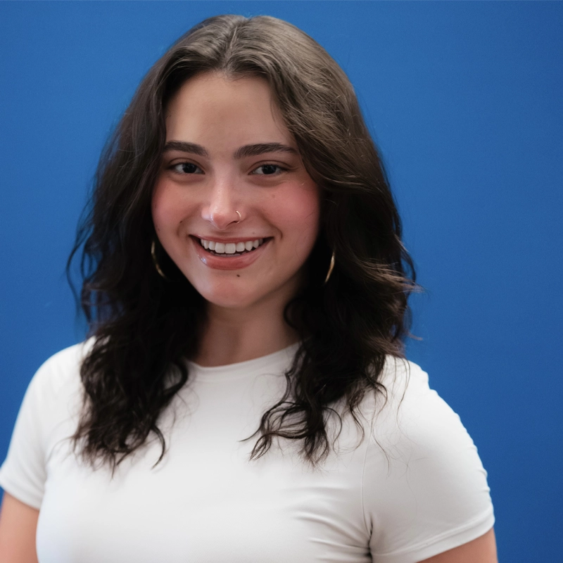 Caroline Cohen, a young woman with wavy dark brown hair and gold hoop earrings, smiles warmly. She wears a fitted white short-sleeve top and stands confidently in front of a solid blue background, her expression friendly and approachable.