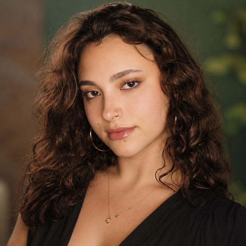 A young woman with long, curly brown hair, wearing a black top, gold hoop earrings, and a delicate gold necklace with small charms, poses slightly smiling against a softly blurred, green and brown background—Caroline Cohen exudes effortless elegance in this portrait.
