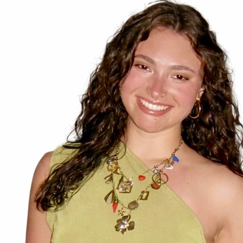 Caroline Cohen, a smiling woman with long, curly brown hair, wears a one-shoulder light green top and a colorful statement necklace. She has hoop earrings and stands against a plain white background.