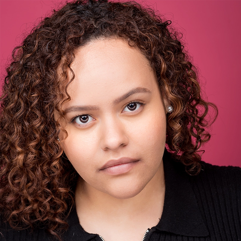 Jenna Willet, with medium brown skin and curly brown hair, stares directly at the camera with a neutral expression. She wears pearl stud earrings and a black top against a solid pink background.
