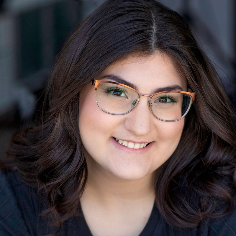 A young woman with wavy, shoulder-length dark hair smiles at the camera. She wears clear-rimmed glasses, a dark top, and has natural makeup. The background is softly blurred, focusing attention on her friendly expression.