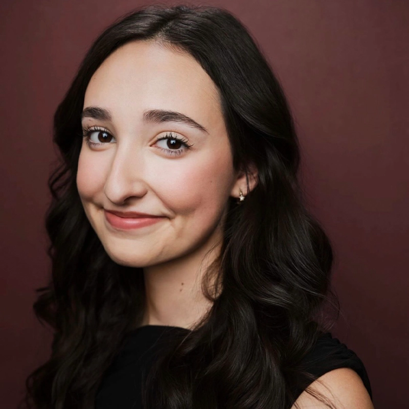 Young woman, Malia Daniels, with long, wavy dark hair and fair skin smiles softly, looking slightly left of the camera. She wears a black top and small earrings, set against a maroon background. The lighting highlights her natural makeup and gentle expression.