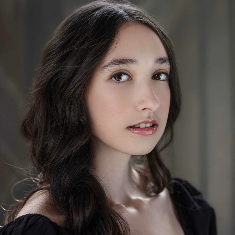 Malia Daniels, a young woman with long, wavy dark brown hair and fair skin, gazes softly at the camera. She wears a black top with off-the-shoulder sleeves and natural makeup. The background is softly blurred with neutral tones.
