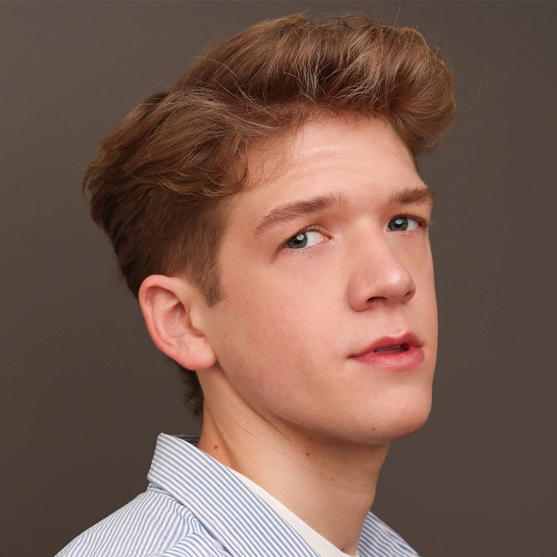 Lazarus Rashid, a young person with light skin and short, wavy brown hair, looks slightly over their shoulder at the camera. They wear a light blue and white striped collared shirt and have a neutral, thoughtful expression against a plain brown background.