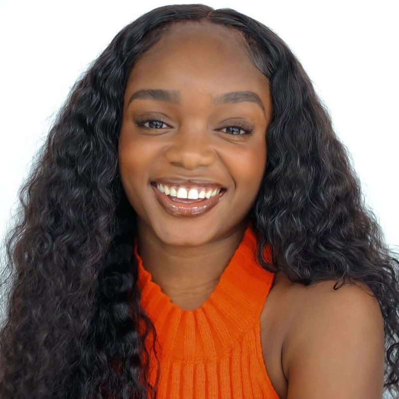 Ary'ana Neveah Davis, a young woman with long, wavy black hair, smiles brightly at the camera. She wears an orange ribbed sleeveless top, and her warm skin tone stands out against the plain white background, highlighting her cheerful expression.