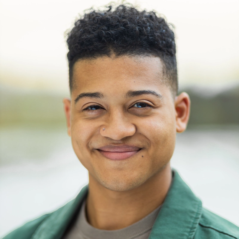 Adrian DeLeon, a young person with short curly hair and medium skin tone, smiles gently at the camera. They have a nose piercing and wear a green jacket over a beige shirt. The softly blurred background draws focus to their face.