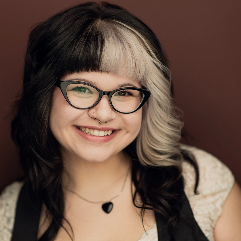 A smiling person with wavy black hair featuring a prominent white streak and bangs, wearing black cat-eye glasses, a nose ring, and a heart-shaped necklace. The background is solid brown, and they are dressed in a light top with dark straps.