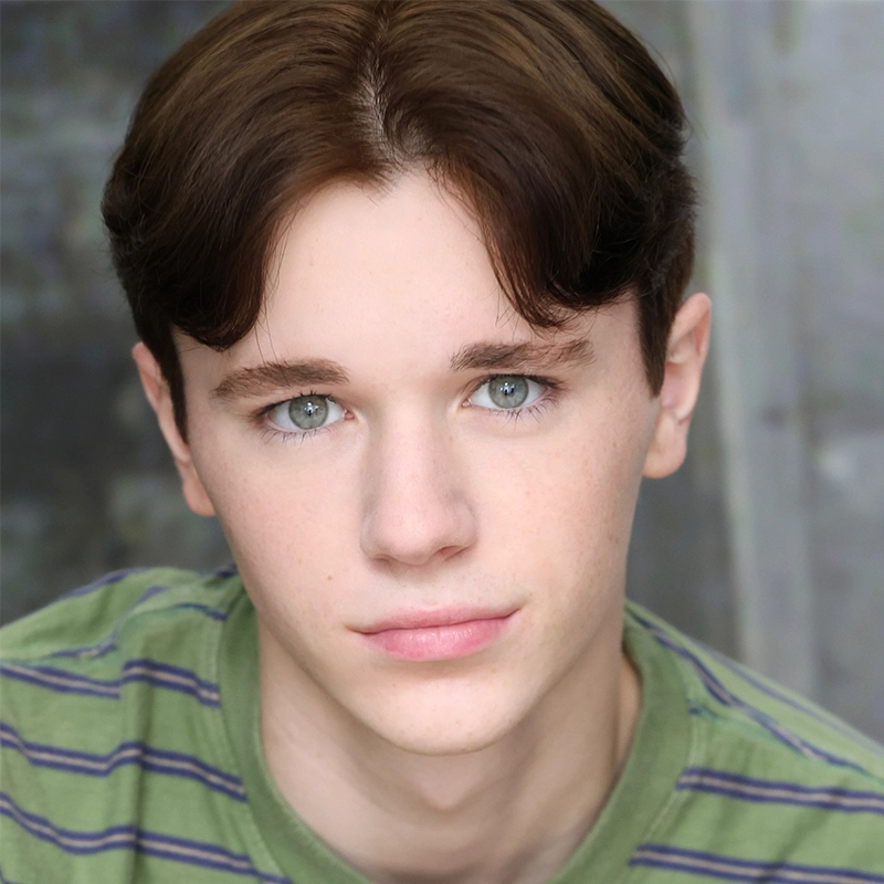 Micah Wolfensohn, a young person with fair skin, light eyes, and short brown hair styled neatly with a center part, gazes directly at the camera. They wear a green shirt with thin blue stripes against a soft, grayish blurred background.