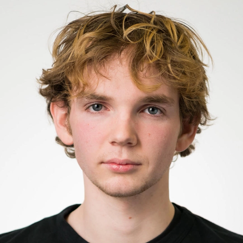 Lexis Trechak, a young person with fair skin, tousled light brown hair, and blue eyes, looks directly at the camera with a neutral expression. They are wearing a black shirt against a plain, light background.