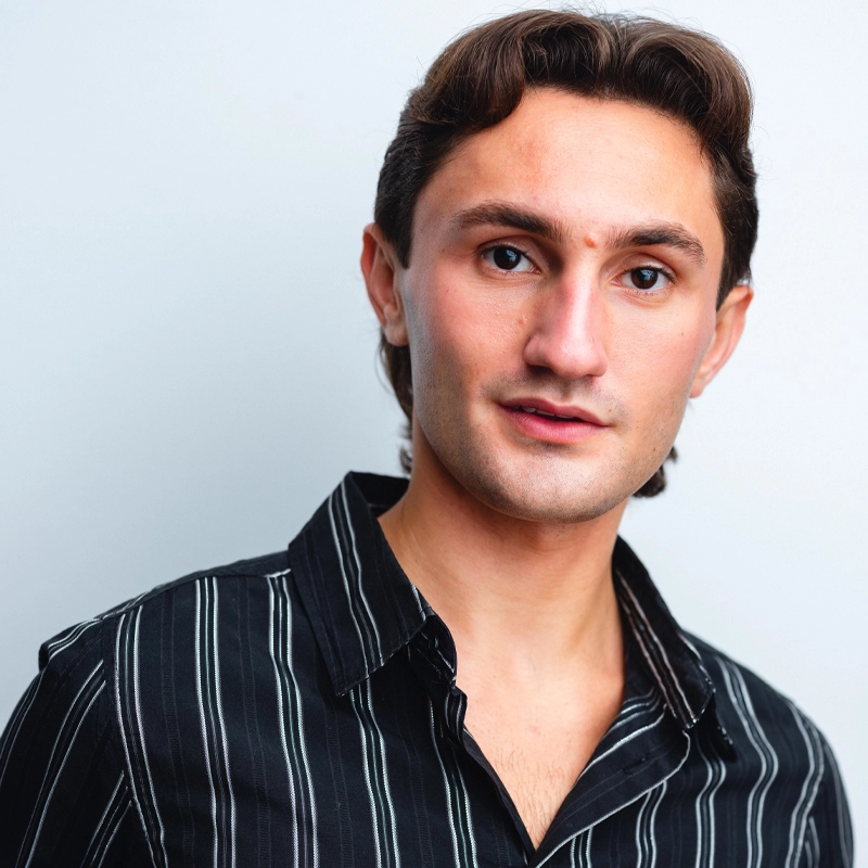 A young man with wavy brown hair wearing a black shirt with white vertical stripes stands against a light gray background. He has light skin, brown eyes, and a slight smile, looking directly at the camera.
