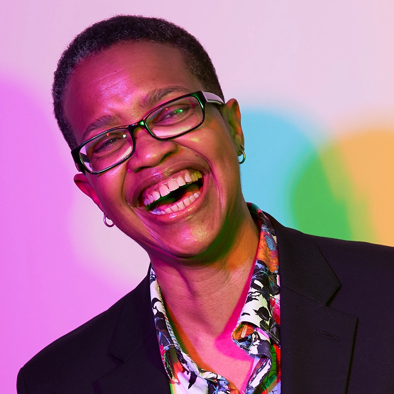Aron Marchak, with short hair, glasses, and small hoop earrings, smiles broadly in a black blazer over a colorful patterned shirt. The white background pops with vibrant pink, green, and yellow lighting effects.