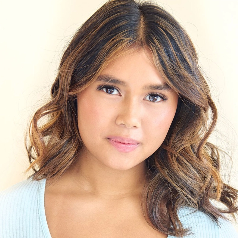 A young woman with wavy, shoulder-length brown hair with blonde highlights looks at the camera. She wears a light blue top and has a calm, neutral expression. The soft beige background creates a bright, gentle atmosphere—portrait by Micah Wolfensohn.