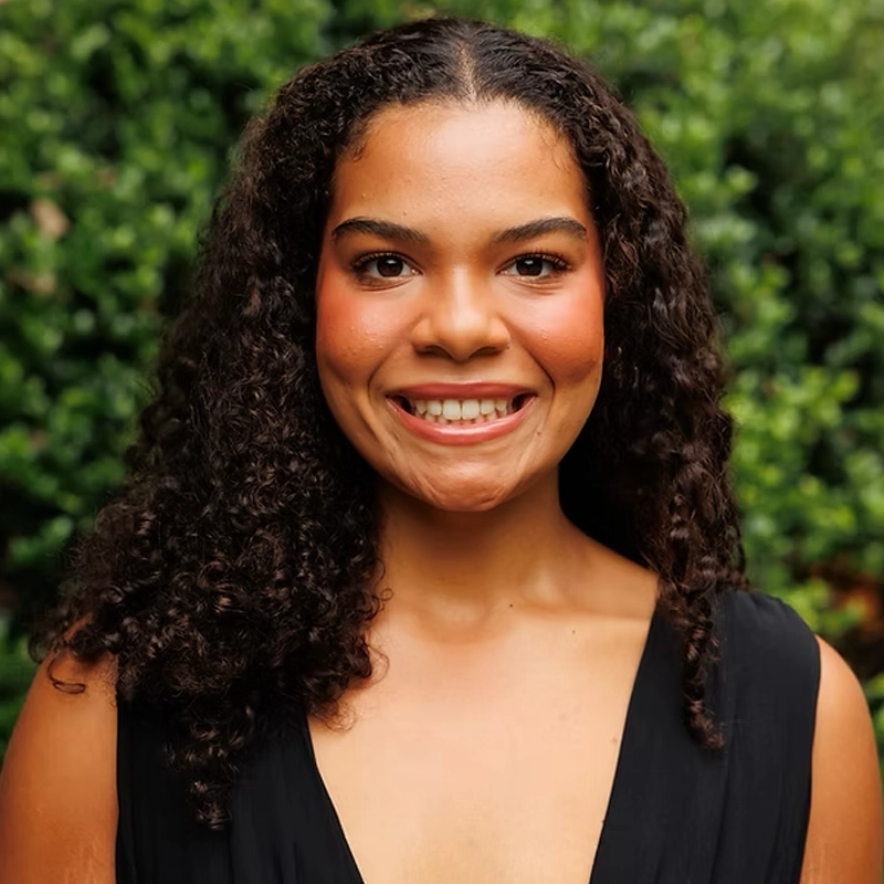 Graci Doster, a woman with curly dark hair and medium brown skin, smiles at the camera in a sleeveless black top. The blurred greenery in the background suggests an inviting outdoor setting.