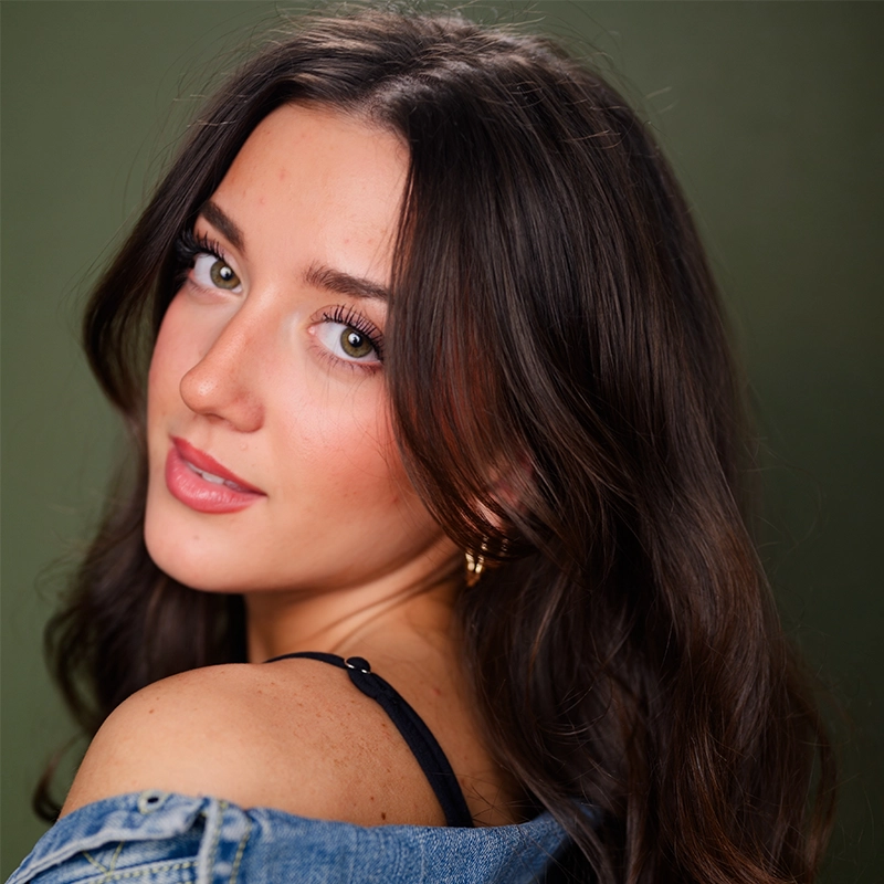A young woman with long wavy dark hair looks over her shoulder at the camera. She wears a denim jacket draped off one shoulder, exposing a black strap. Her makeup is natural, and the background is a soft, muted green.
