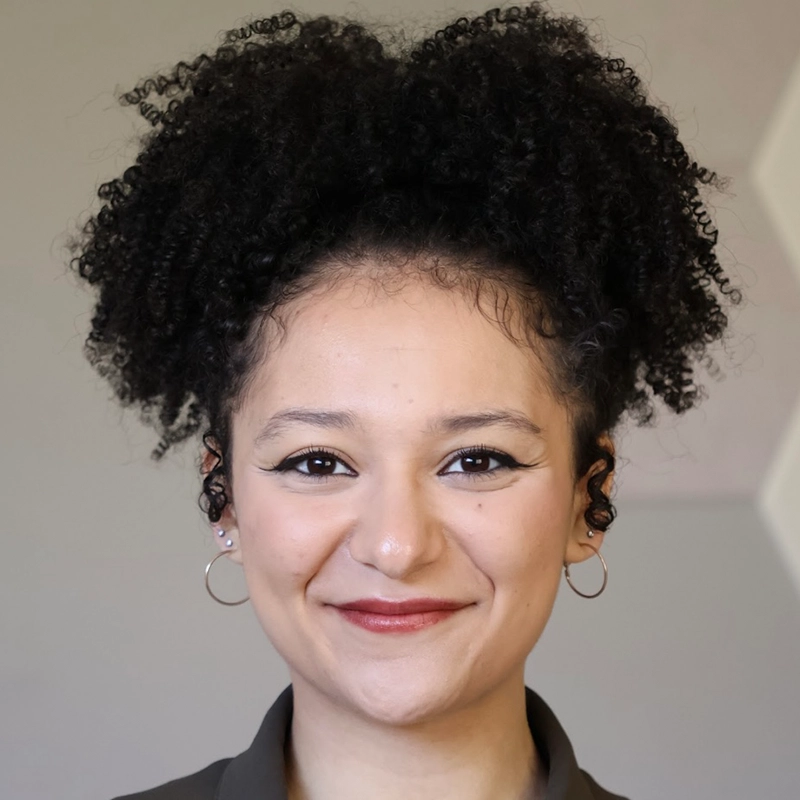Addison Zacharias, a young woman with curly dark hair styled in a high puff, smiles slightly. She wears hoop earrings, subtle makeup with winged eyeliner, and a dark collared shirt. The softly blurred background is beige and light gray.
