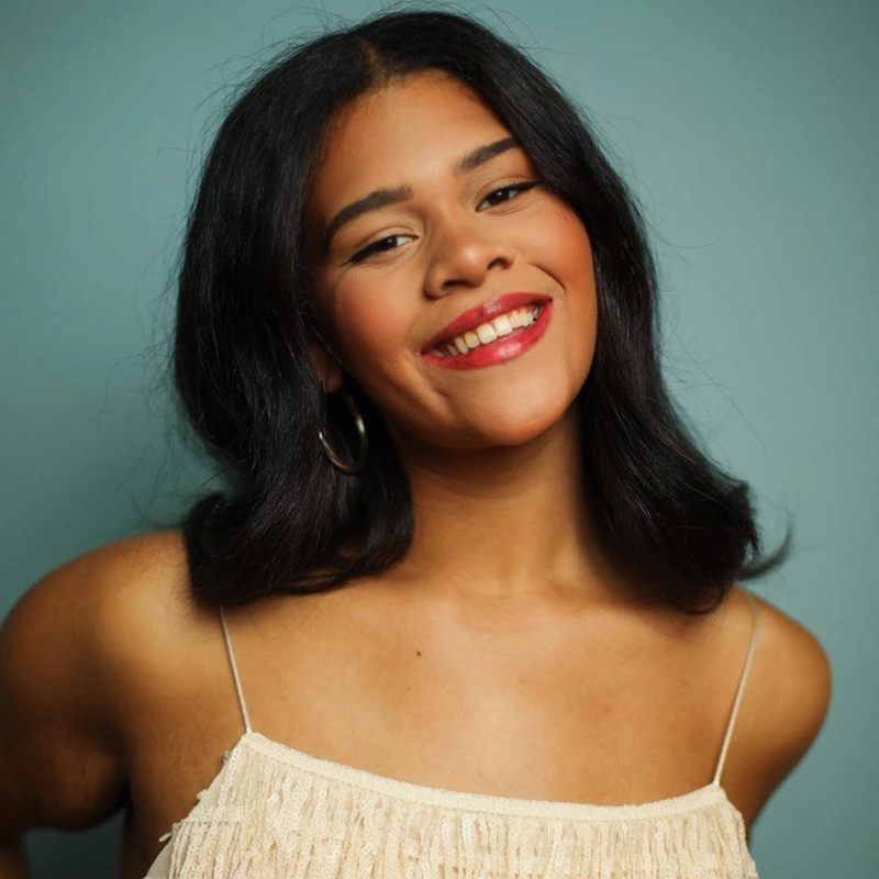 Lexi Einsenhart, a young woman with medium skin tone and shoulder-length dark hair, smiles warmly at the camera. She wears hoop earrings, red lipstick, and a cream-colored, thin-strapped top against a soft teal blue background.
