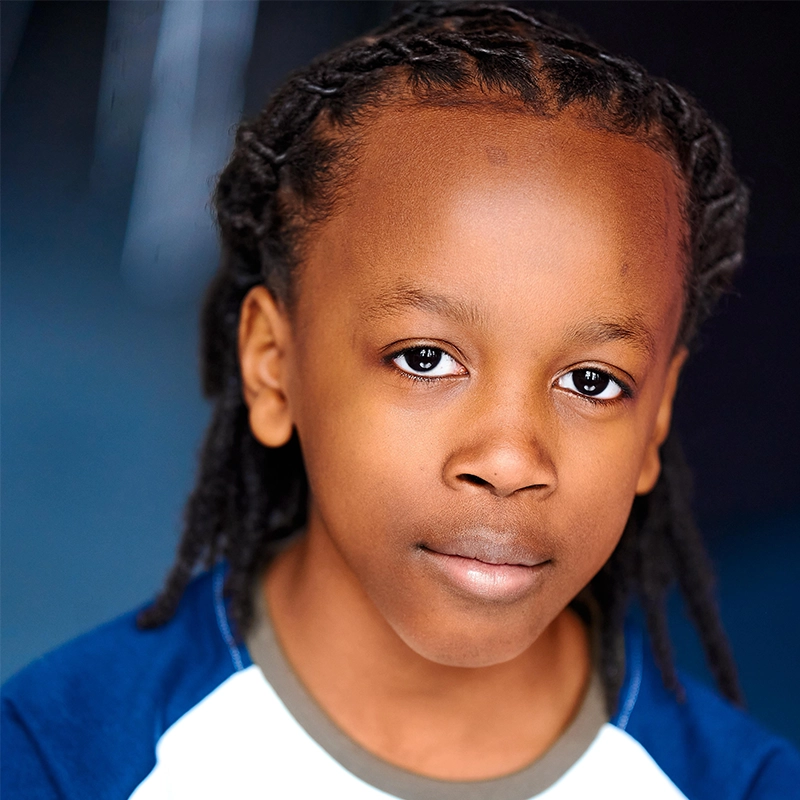 A young Black boy with neat, shoulder-length braids gazes calmly at the camera. Wearing a blue and white shirt, he has a slight, relaxed smile. The softly blurred background keeps the focus on his expressive face—reminiscent of Kenneth E. Johnson Jr.'s portraits.