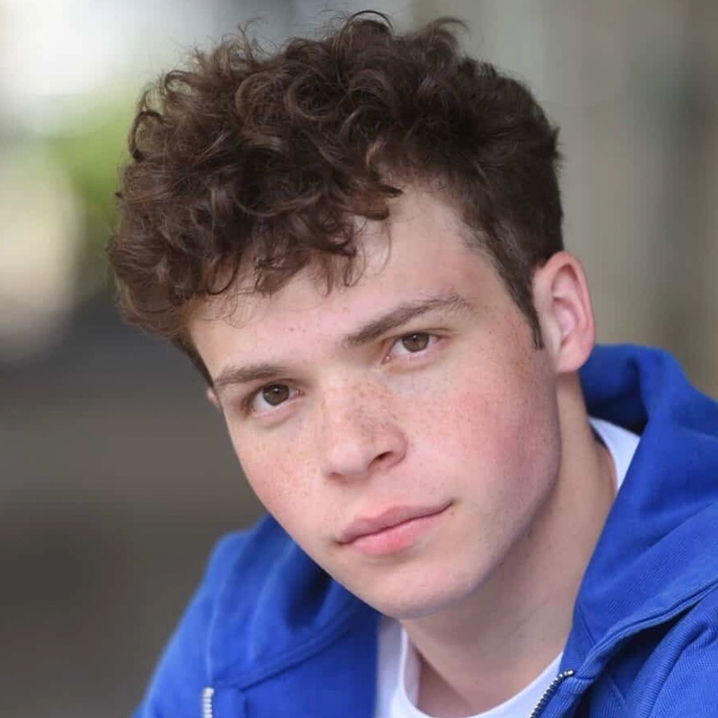 Micah Wolfensohn, a young man with short, curly brown hair and fair skin, looks at the camera with a neutral expression. He is wearing a bright blue jacket over a white shirt, and the softly blurred background glows with natural light.
